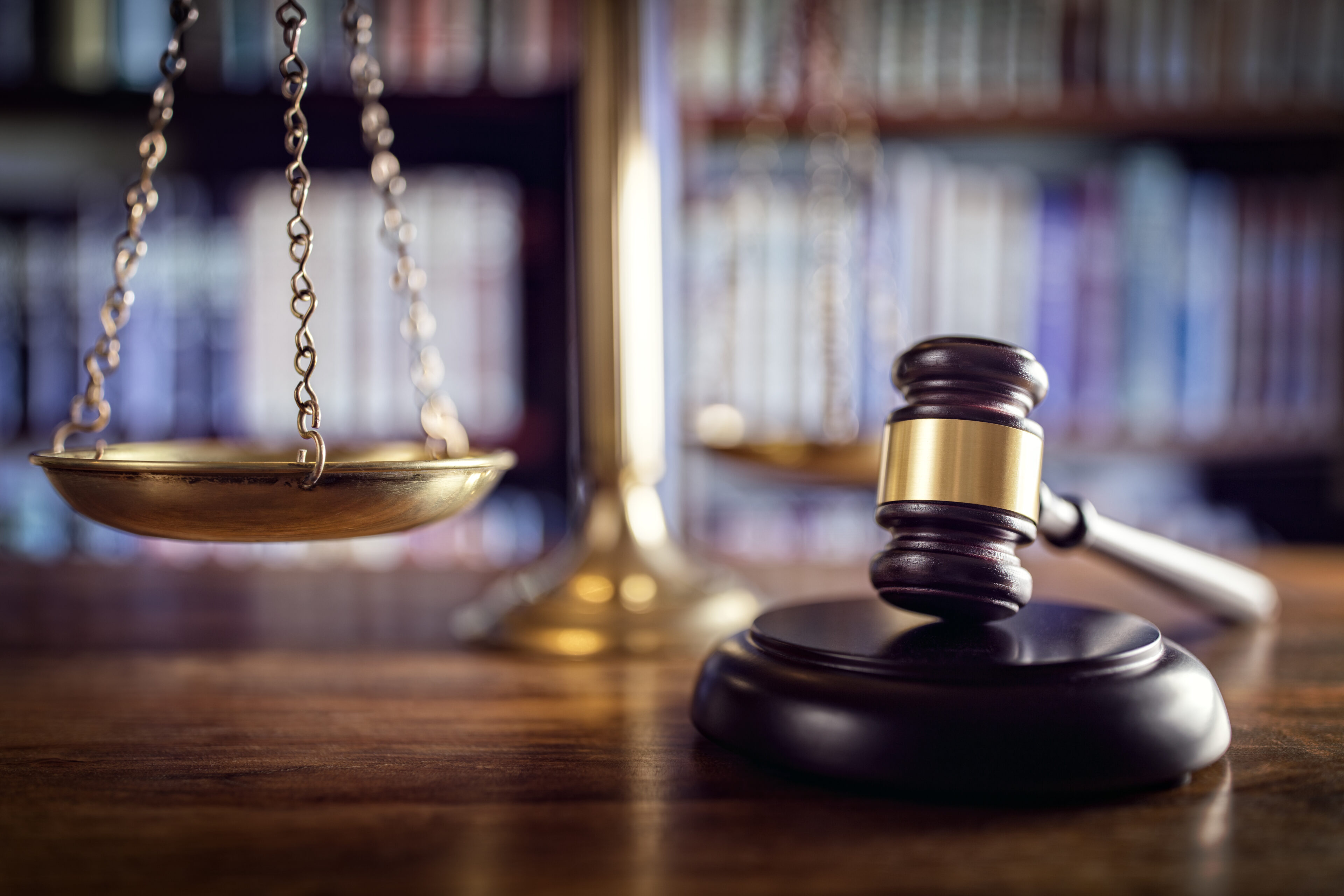 Wooden judge's gavel resting on a desk with a blurred balance scale and bookshelves in the background, symbolizing legal proceedings, justice, and malpractice litigation.