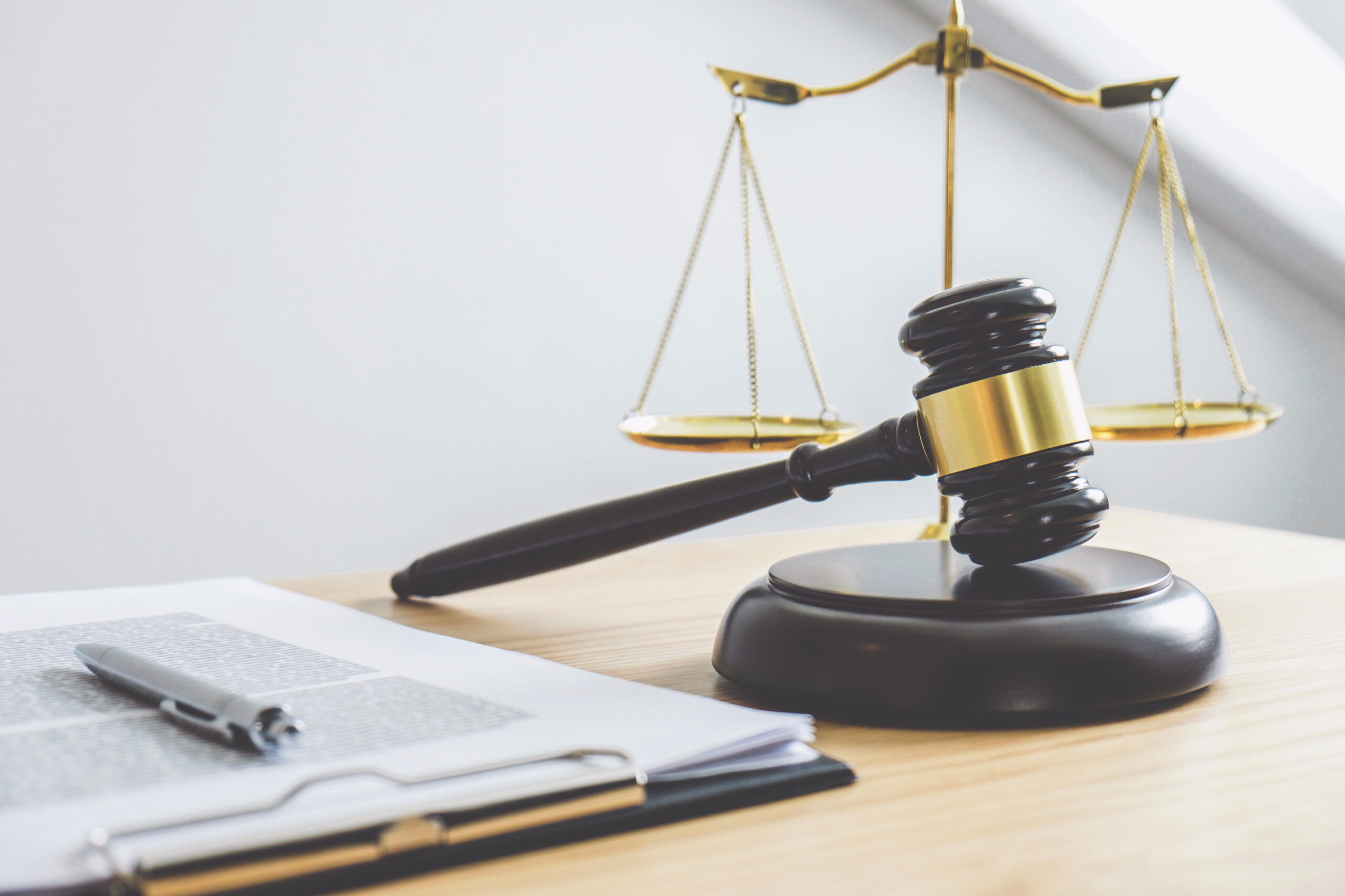 Wooden gavel resting on a sound block beside a legal notepad and pen, with a brass balance scale in the background, symbolizing law, justice, litigation, and legal proceedings.