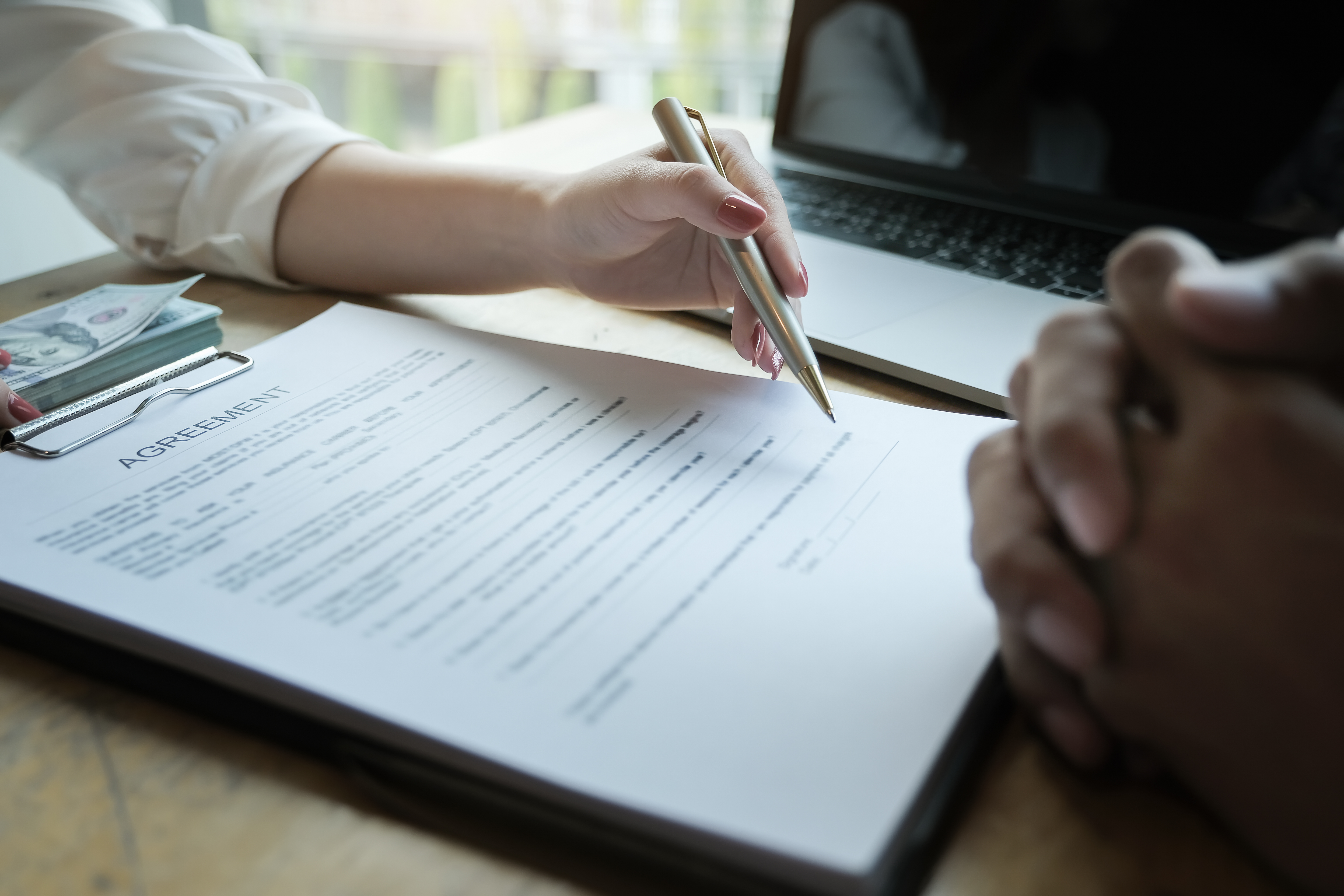 Person reviewing and signing a document on a clipboard beside a laptop during a meeting.