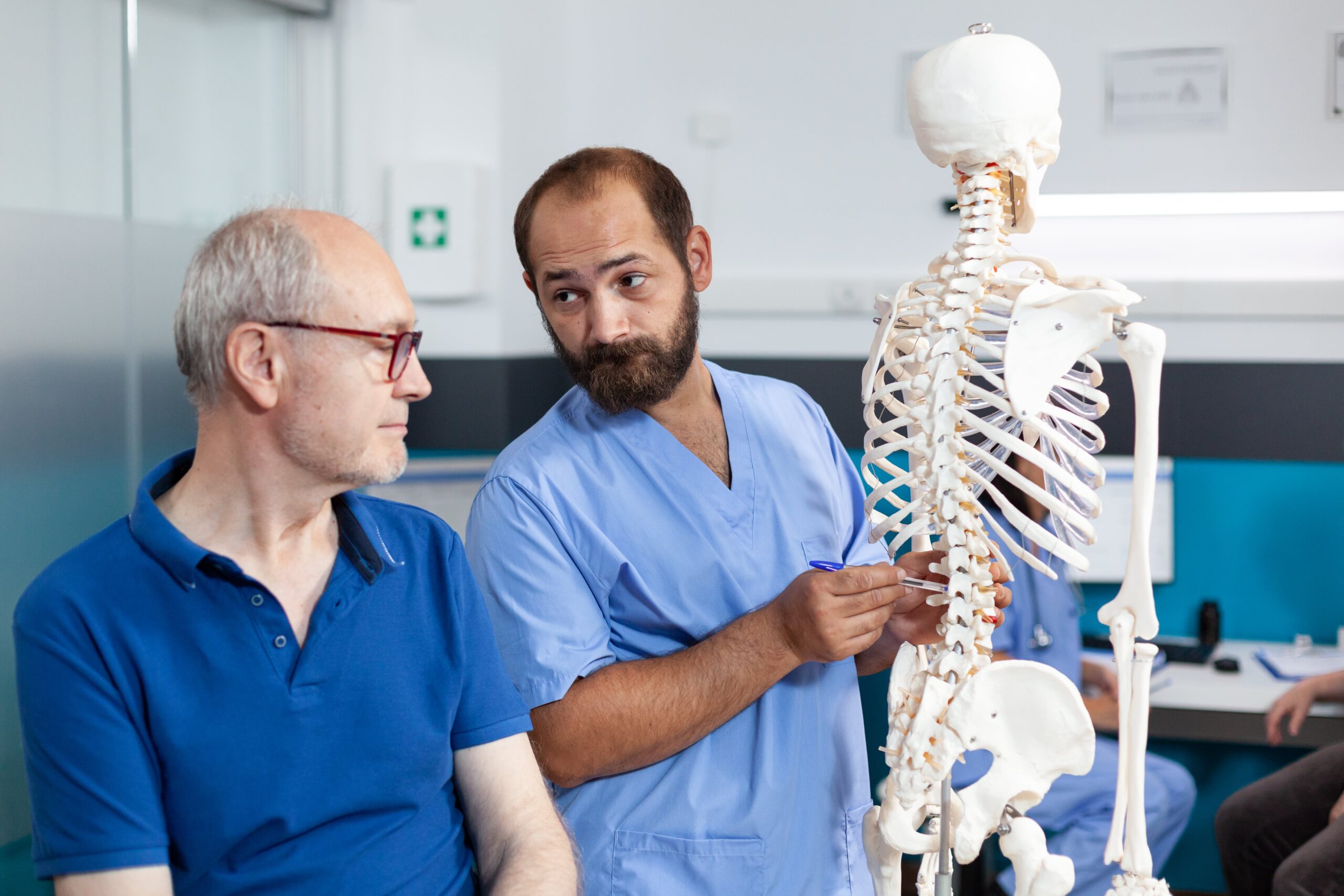 Healthcare professional explaining spinal anatomy using a skeleton model to an older patient in a clinic setting.
