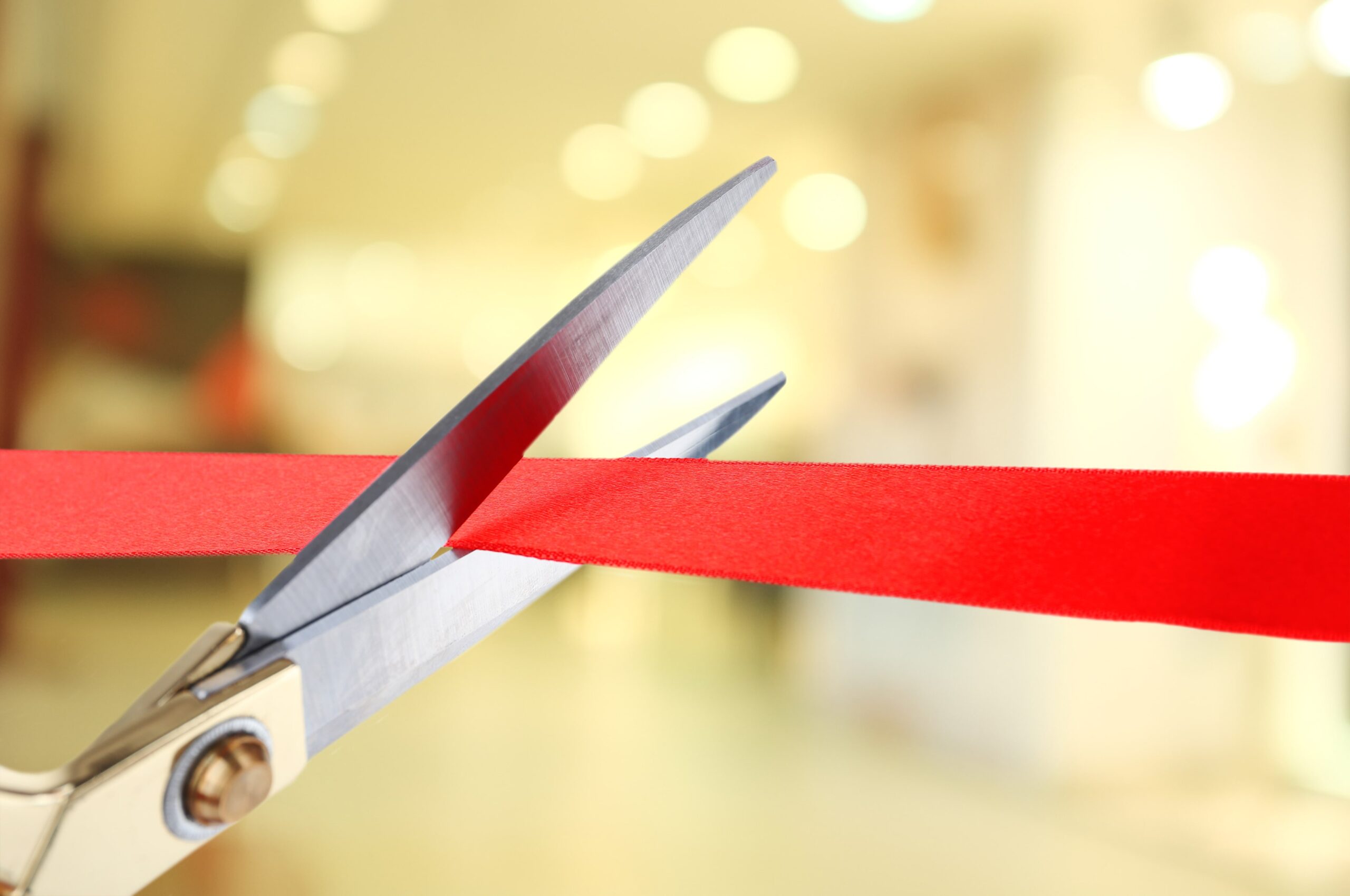 Close-up of silver scissors cutting a red ribbon during a ribbon-cutting ceremony, with a softly blurred indoor background.