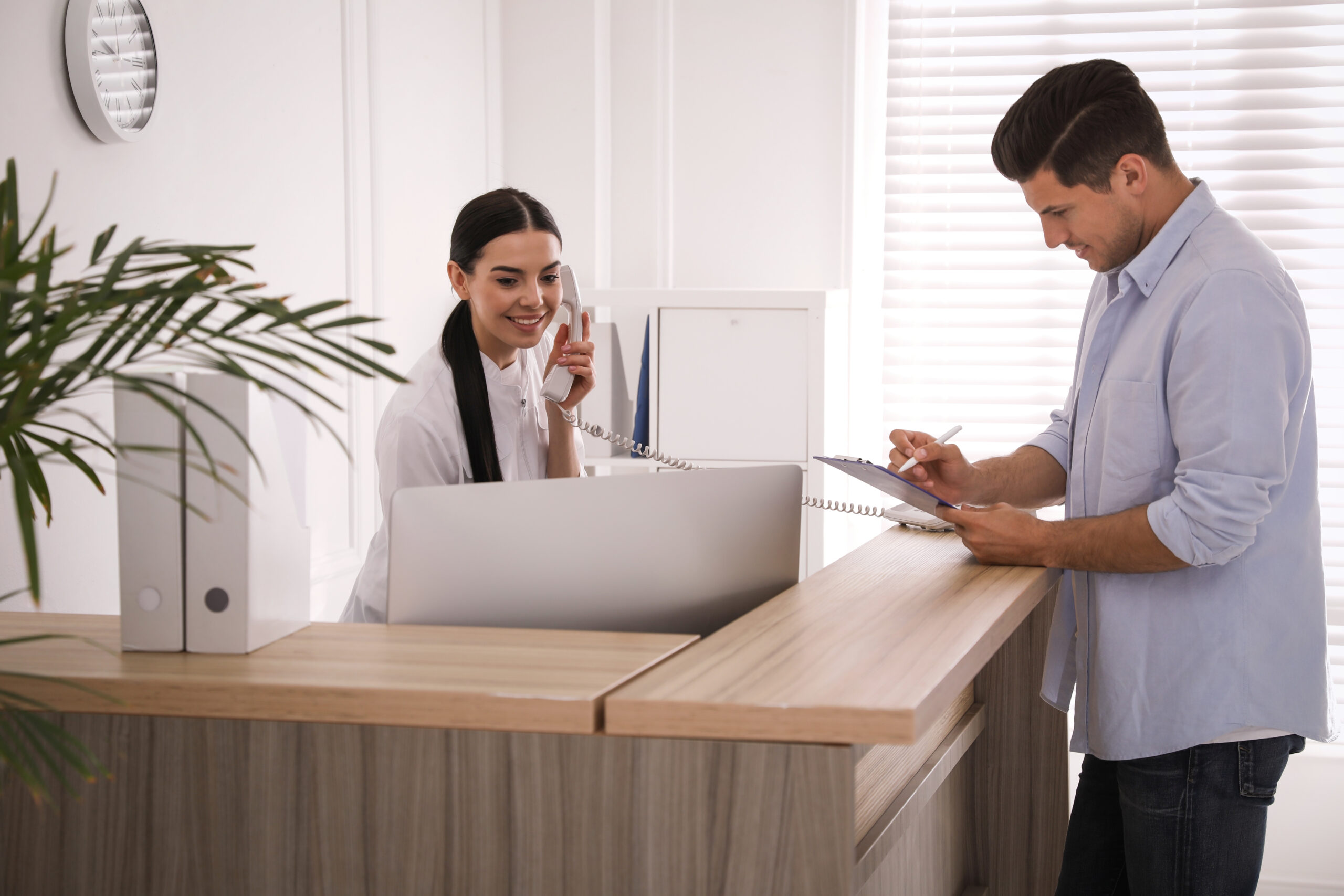 Person standing at a reception desk using a smartphone while another person works at a computer behind the counter.