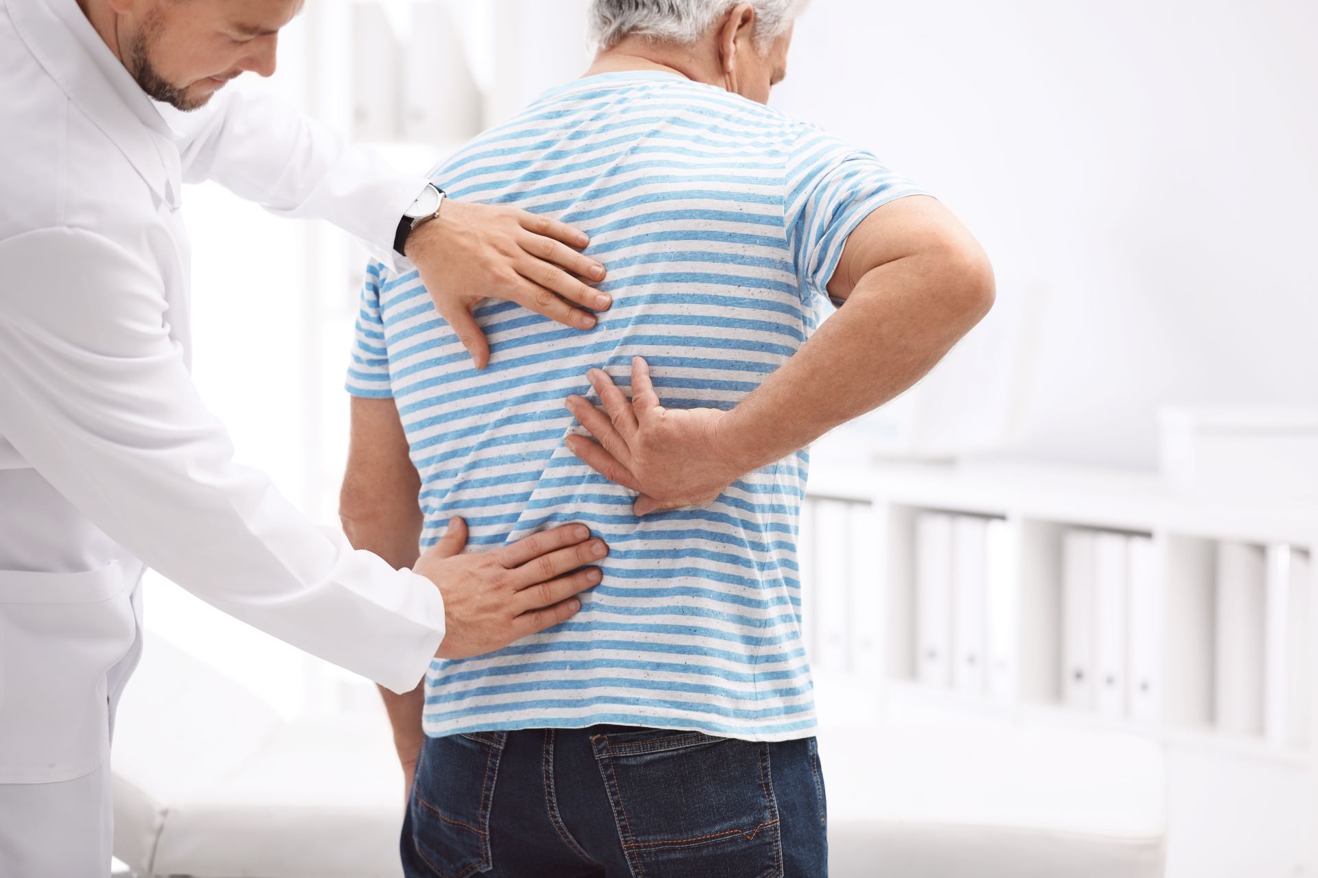 Chiropractor examining an older patient’s back while the patient places a hand on the lower back in a clinic setting.