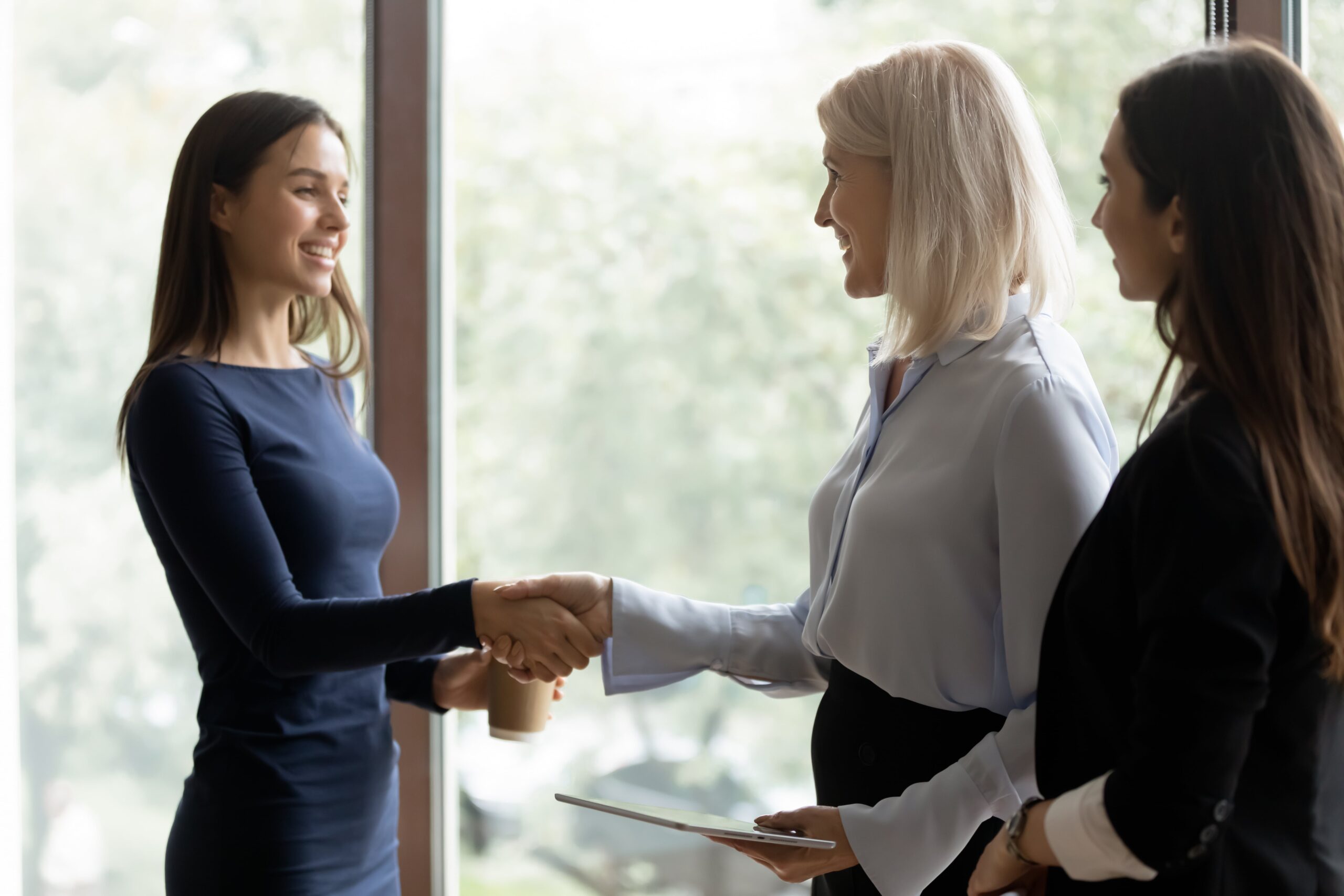 Three professionals shaking hands during a business meeting while two colleagues stand nearby holding documents.