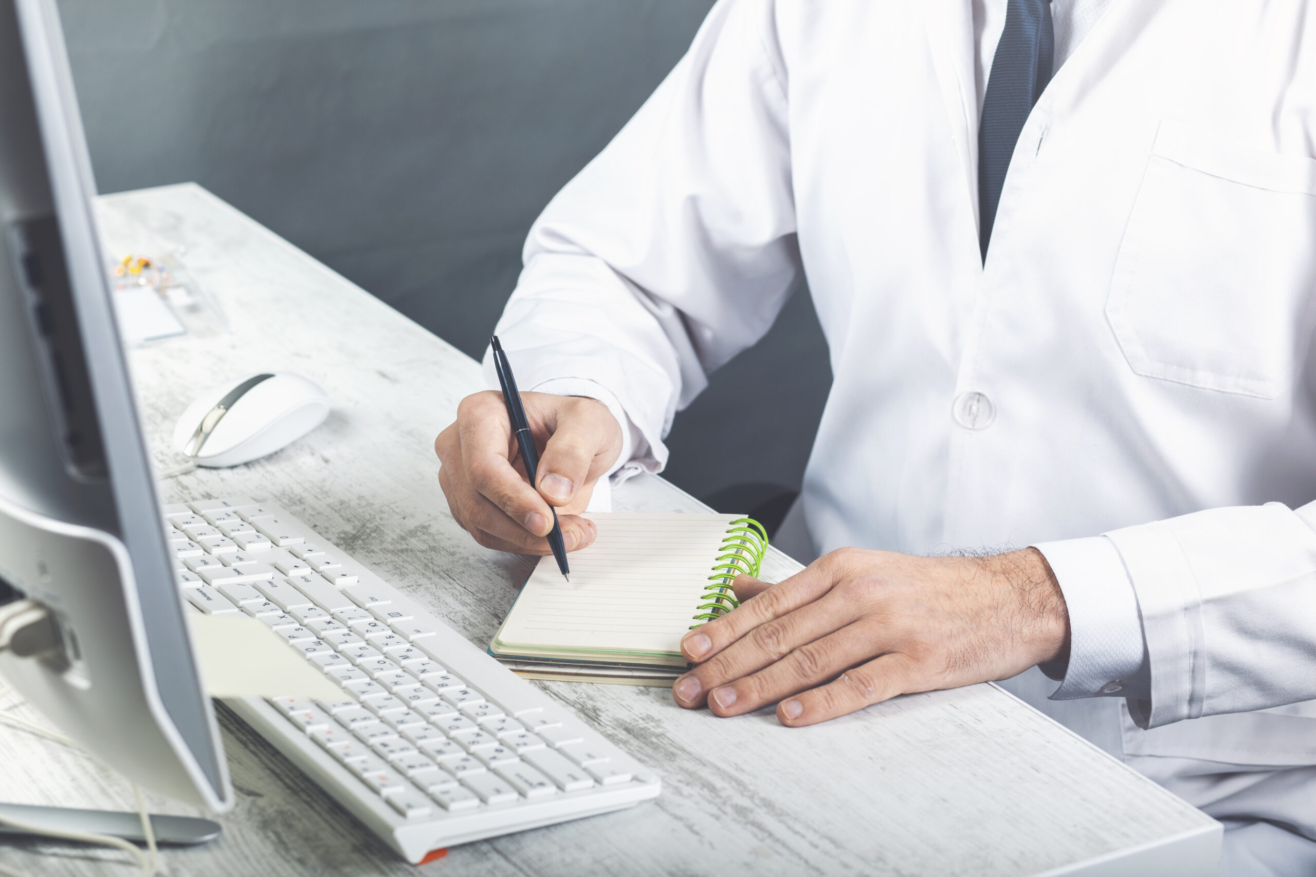 Chiropractor in a white coat writing notes on a clipboard beside a computer keyboard at a desk.
