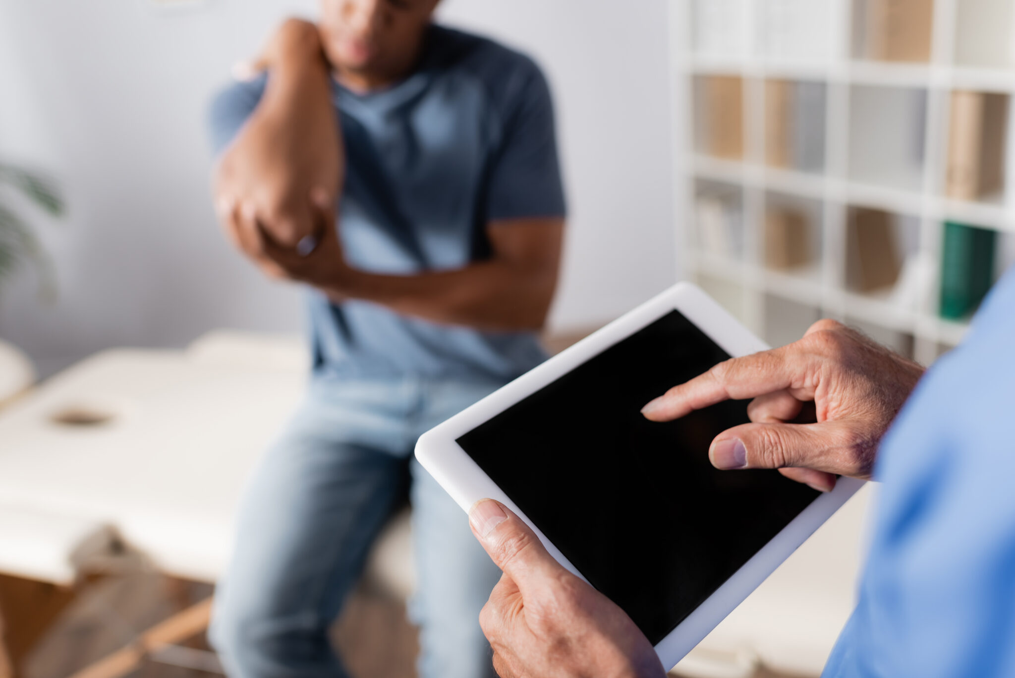Healthcare professional holding a tablet while consulting with a patient seated in an exam room.