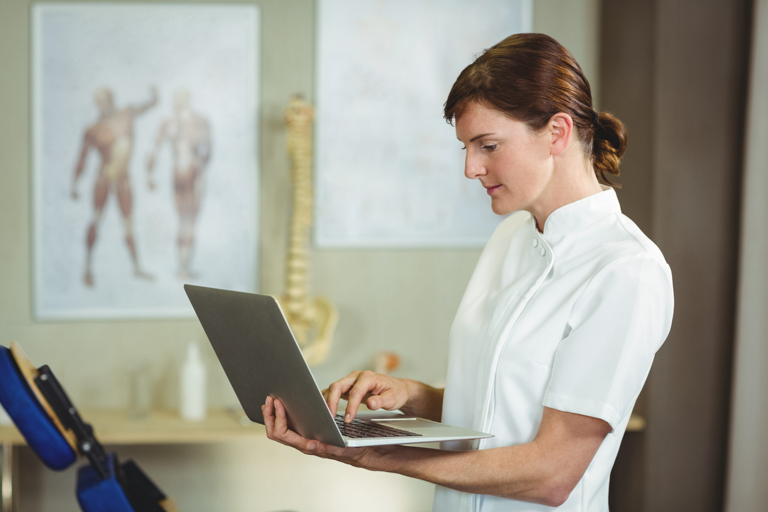 Healthcare professional in a white coat using a laptop at a desk, with an anatomical chart visible in the background.